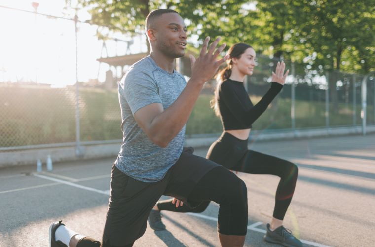 Two people are exercising outdoors, performing lunges on a paved surface. Sunlight filters through the trees in the background as they focus on strengthening their legs to prevent knee injuries. They are wearing athletic clothing.