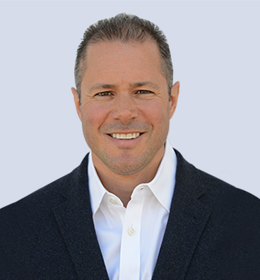 A man in a dark blazer and white shirt smiles at the camera against a light background, exuding positivity despite recovering from a knee injury.