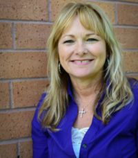 A smiling woman with long blonde hair stands against a brick wall, wearing a purple shirt and a cross necklace, her confidence undeterred by a recent shoulder injury.