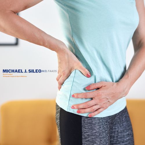 Person standing indoors, wearing a light blue shirt and dark pants, holding their right hip and lower back area with both hands, possibly indicating discomfort or pain.
