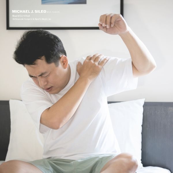 A man sitting on a bed holds his shoulder and appears to be in pain, as if experiencing discomfort or an injury.