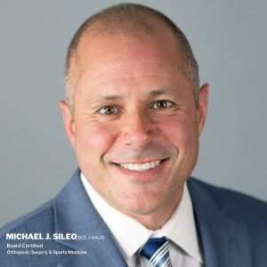 A man in a blue suit and striped tie smiles at the camera against a plain gray background. Text in the lower left corner displays his name and credentials. A man in a blue suit and striped tie smiles at the camera against a plain gray background Text in the lower left corner displays his name and credentials