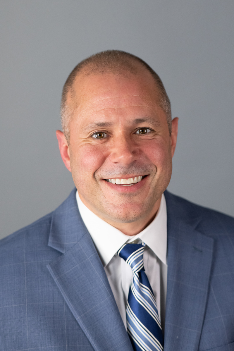 A man in a blue suit, white shirt, and striped tie smiles at the camera against a plain gray background.