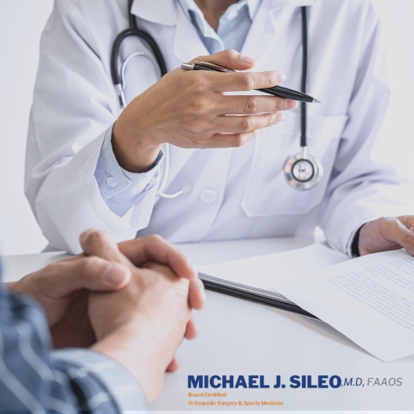 best orthopedic knee surgeons on long island dr.michael sileo (1) A doctor in a white coat, known among the Best Orthopedic Knee Surgeons on Long Island, holds a pen while discussing paperwork with a patient across a desk; a stethoscope hangs around the doctor's neck.
