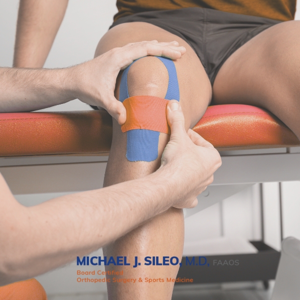 sileo sports medicine doctor near melville A Sports Medicine Doctor Near Melville applies colorful athletic tape to another person's knee while seated on an examination table.
