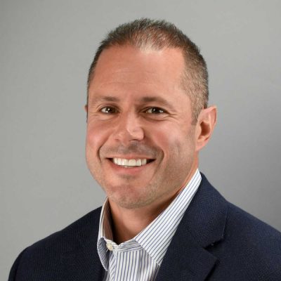 A man in a dark blazer and striped shirt smiles at the camera against a plain gray background.