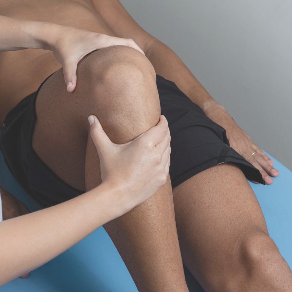A sports medicine physiotherapist holds and examines a patient's bent knee while the patient lies on a blue examination table. A sports medicine physiotherapist holds and examines a patient's bent knee while the patient lies on a blue examination table