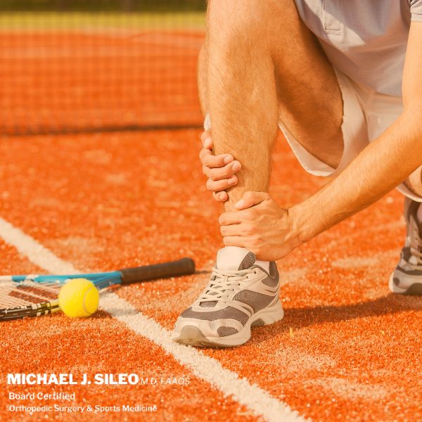 A tennis player on a clay court holds his ankle in pain, with a racket and tennis ball nearby. Text in the corner reads "Michael J. Sileo, M.D., Sports Medicine East Setauket NY, Orthopedic Surgery & Sports Medicine. Tennis player holding an injured ankle on the court — sports medicine care in East Setauket, NY