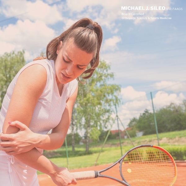Female tennis player on an outdoor court holds her right elbow in pain, her racquet resting on the net under a partly cloudy sky—an injury best evaluated by Sports Medicine Commack NY specialists. Sport Injury Dr Michael Sileo Treats